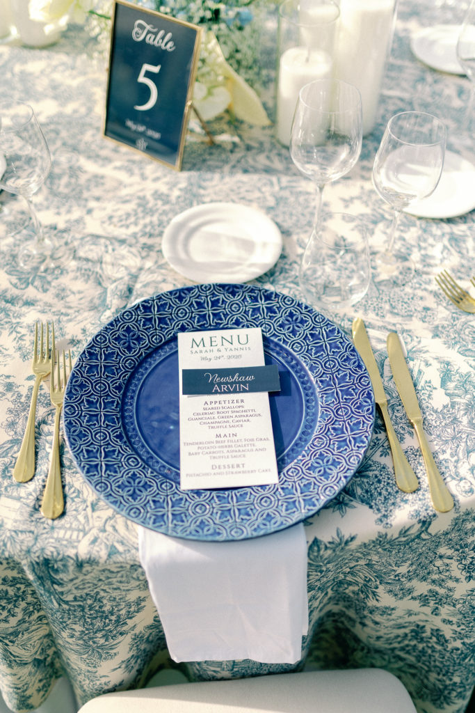 Close-up of a wedding reception table in Athens with blue patterned charger, printed menu, candles, and gold cutlery.