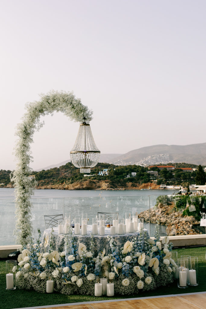 Bride and groom seated at candlelit sweetheart table under a floral chandelier with the sea in the background at a destination wedding in Greece.