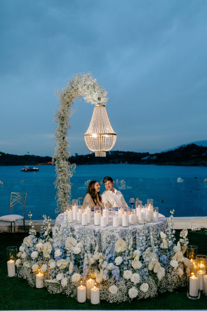 Bride and groom seated at candlelit sweetheart table under a floral chandelier with the sea in the background at a destination wedding in Greece.