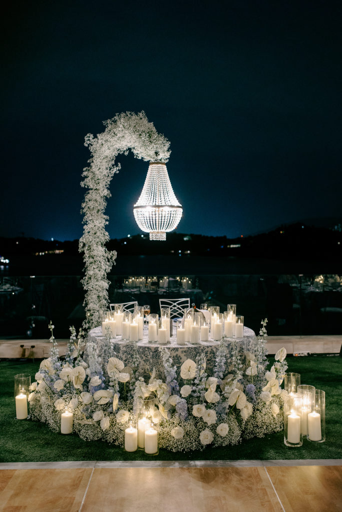 Bride and groom seated at candlelit sweetheart table under a floral chandelier with the sea in the background at a destination wedding in Greece.