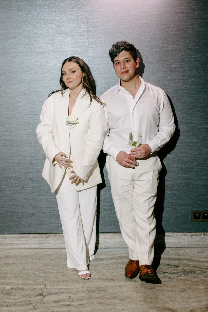 Elegant romantic wedding in Athens - Party -A man and woman dressed in white stand against a gray wall, the woman holds a white rose and the man holds a glass.