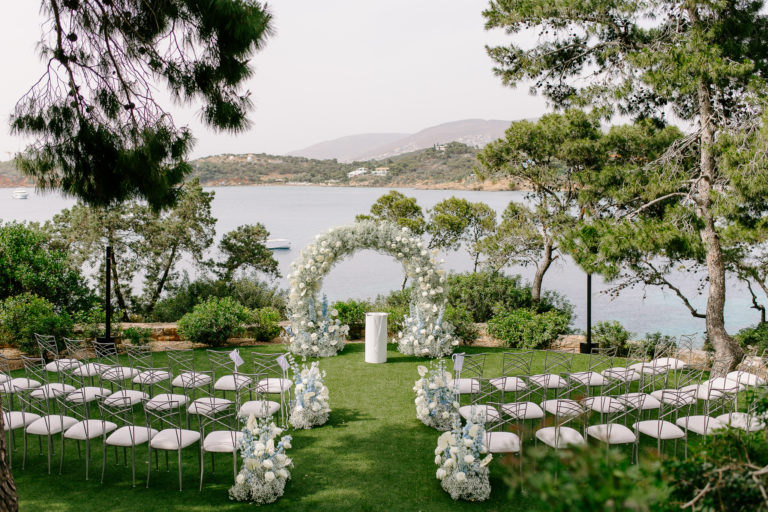 Elegant outdoor wedding ceremony setup in Greece with a white floral arch, chairs facing the sea, and lush greenery overlooking the sea.