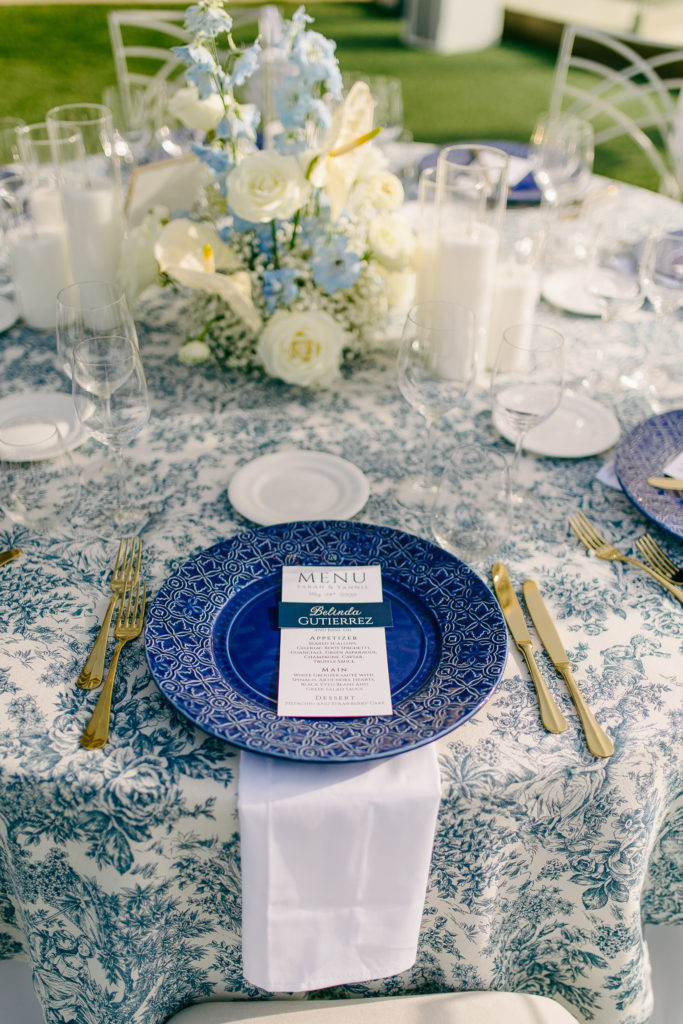 Close-up of a wedding reception table in Athens with blue patterned charger, printed menu, candles, and gold cutlery.