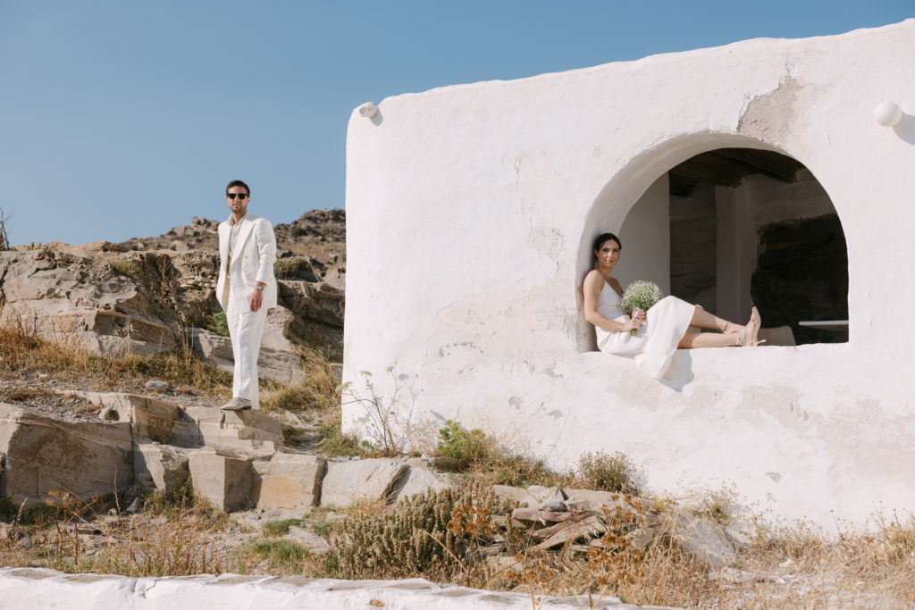 Bride sitting in a rustic white Cycladic arch and groom standing on rocky terrain during a minimalist Paros island wedding photo session.
