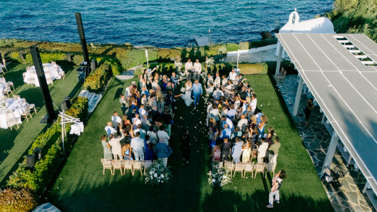 Aerial view of an elegant cliffside wedding ceremony in a garden overlooking the sea on the Athens Riviera, featuring white floral arrangements and guests gathered around the couple.