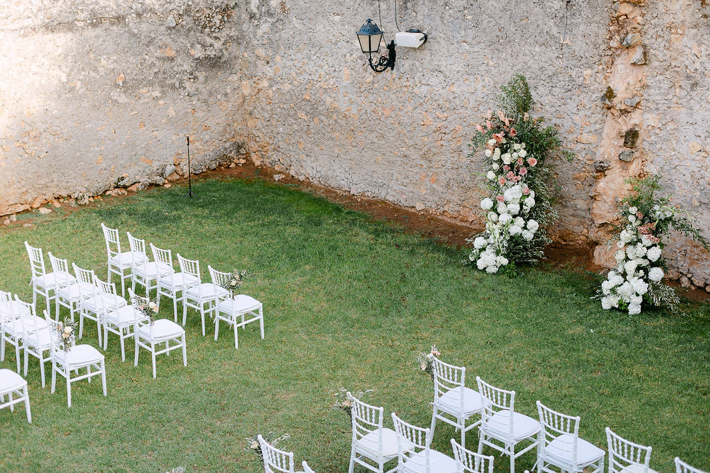 Minimal rustic ceremony setup with white chairs and green floral arrangements against an old stone wall in Crete.