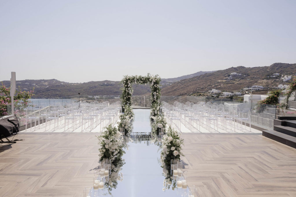 Luxurious Mykonos wedding ceremony with mirrored aisle, white and green florals, and a cliffside sea view.