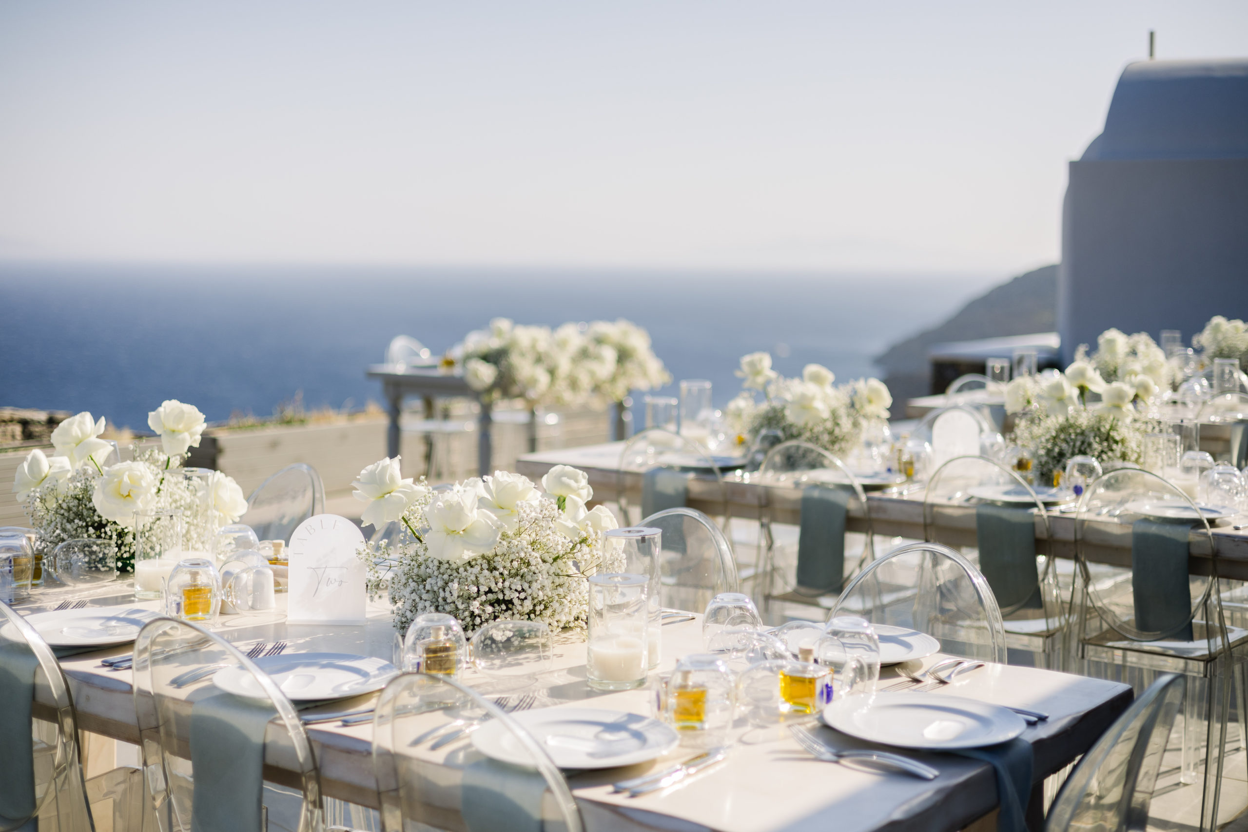 Outdoor wedding reception in Mykonos featuring sea views, minimalist white floral centerpieces, transparent ghost chairs, and elegant table settings under the Greek island sky.