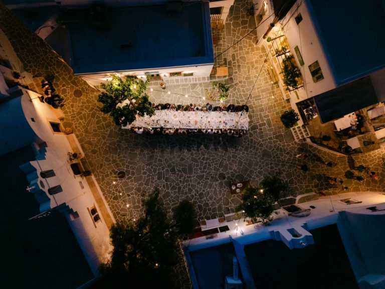 Drone photo of destination wedding dinner in Folegandros village square with long candlelit table and white Cycladic buildings – Greece wedding planner