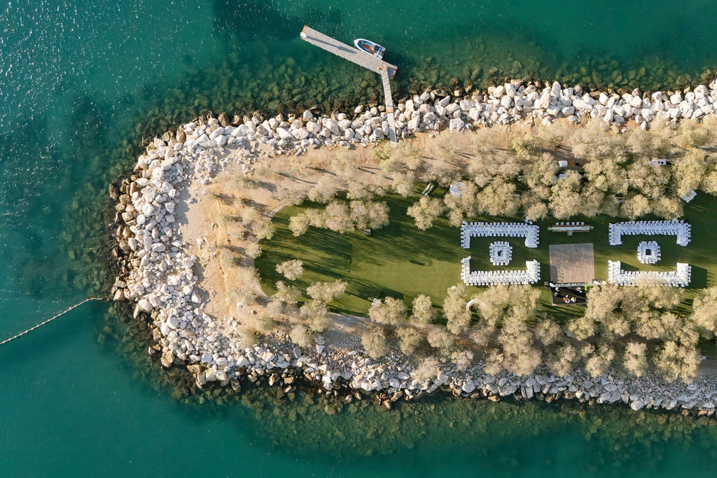 Seaview Wedding on Athens Riviera with White-Purple Florals — aerial view of a coastal lawn ceremony with long tables, white chairs, and abundant white and purple blooms by the turquoise sea.